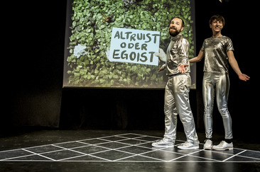 Zwei Schauspieler in silberner Kleidung halten ein Schild mit der Aufschrift Altruist oder Egoist hoch 