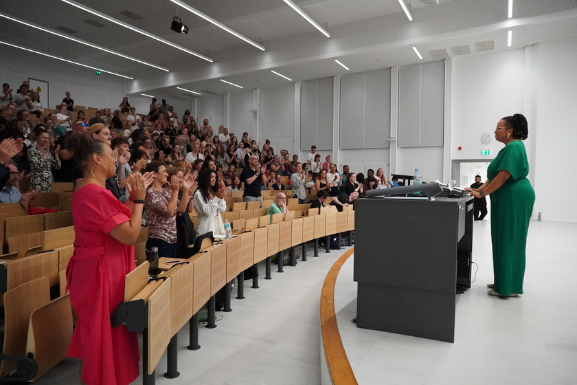 Eine Frau steht in einem grünen Kleid hinter einerm Rednerpult. Man sieht sie von der Seite. Vor ihr, auf der linken Seite des Bildes stehen Menschen, die applaudieren.